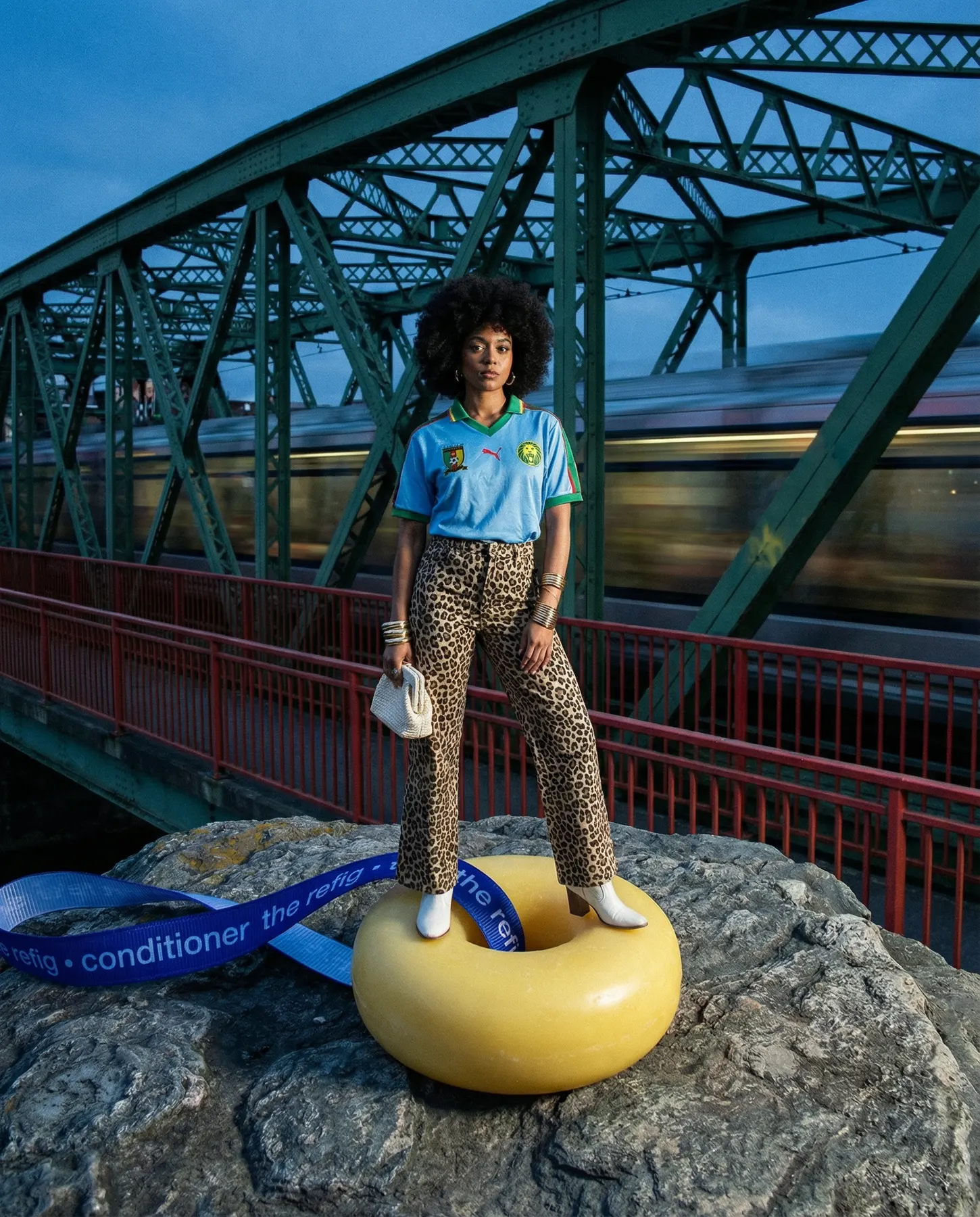 A woman in a blue football shirt and leopard-print trousers stands on a rock by a green steel bridge at dusk, beside a large yellow ring and a blue ribbon printed “the refig conditioner”, with a train blurred behind her.
