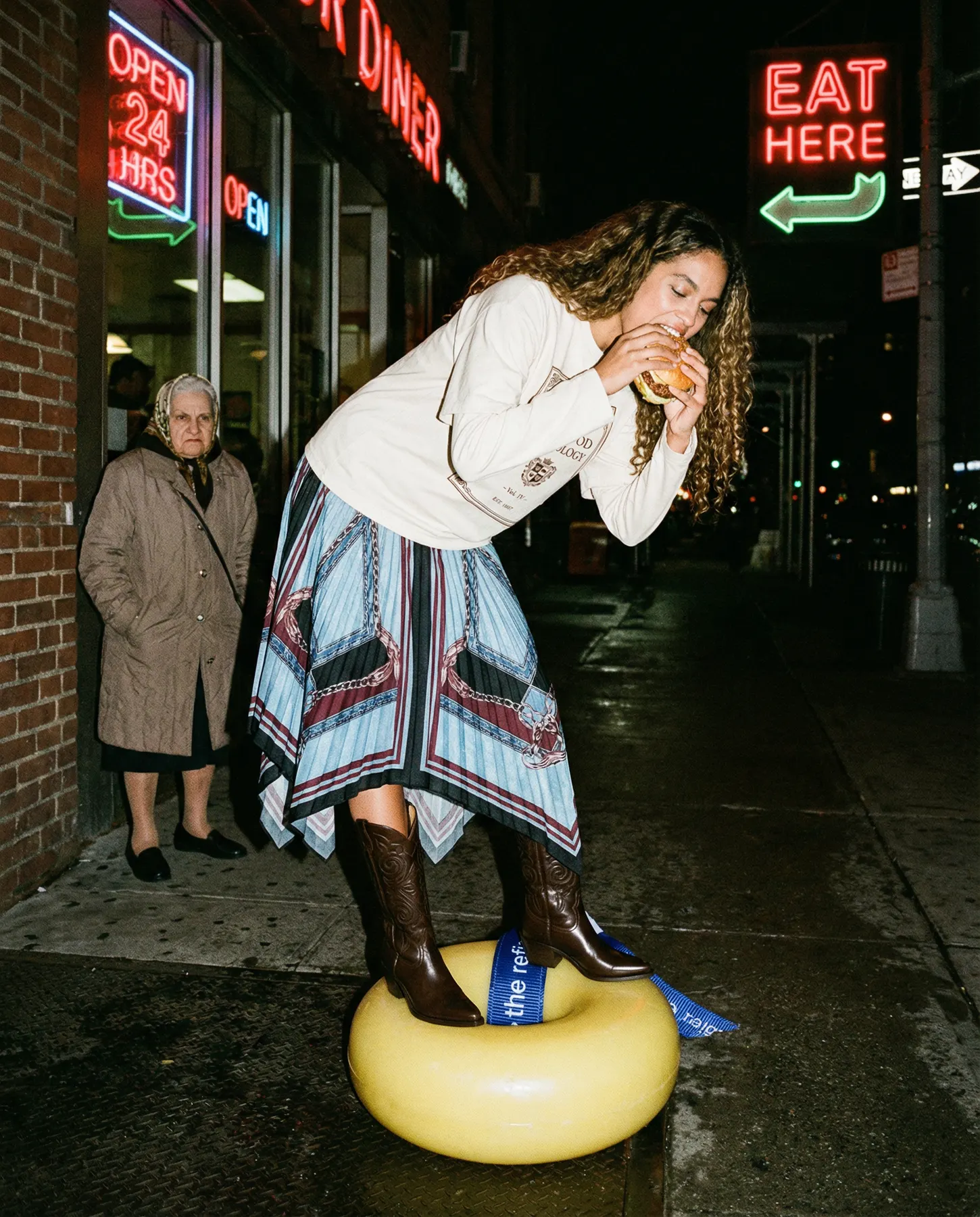 A woman in a cream sweatshirt and a patterned skirt bites into a sandwich on a wet city street at night, one boot on a large yellow ring tied with a blue The Refig ribbon, as an older woman watches from a diner doorway.
